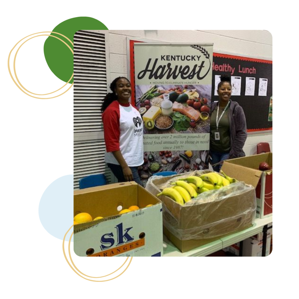 Two women standing in front of a Kentucky Harvest pull-up banner, behind boxes full of fresh fruit.